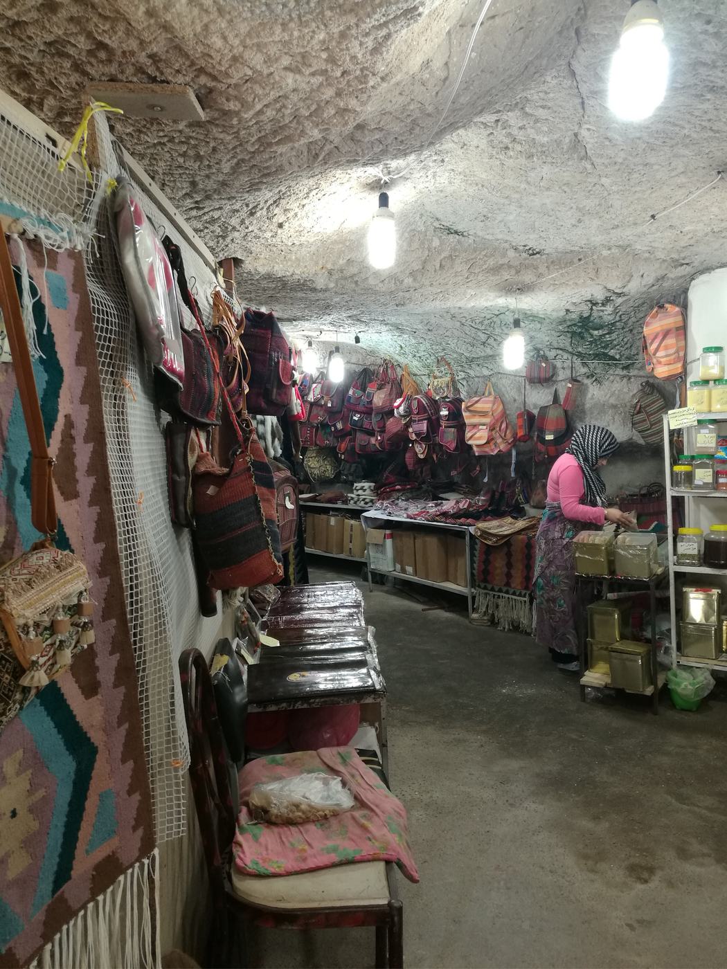 Interior of a small artisan shop carved into stone, displaying colorful woven bags and textiles, with a woman working behind a counter.